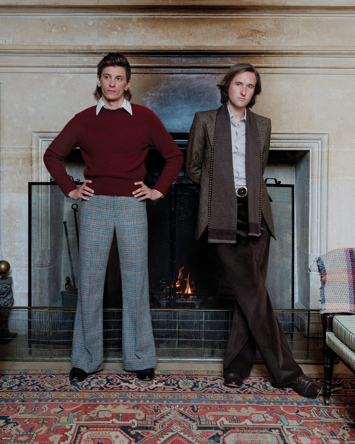 Two men posing in front of a fireplace in a classic interior setting. The man on the left wears a maroon sweater and blue-gray plaid trousers, while the man on the right wears a brown tweed blazer, white shirt, and dark brown trousers.